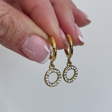 Load image into Gallery viewer, A close-up of a hand holding a pair of gold huggie hoop earrings with dangling circular crystal-studded charms against a plain light gray background.