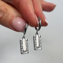 Load image into Gallery viewer, A close-up of a hand holding a pair of silver huggie hoop earrings with rectangular textured dangles against a soft light gray background.